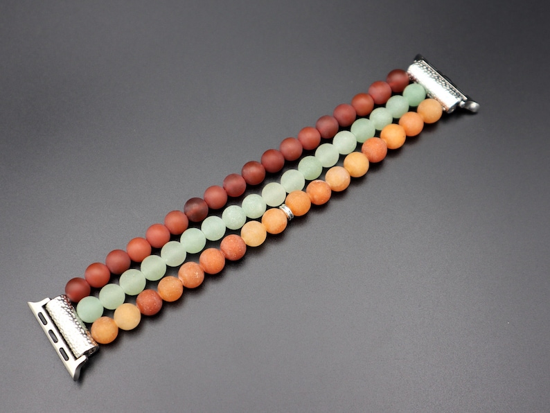May include: A multi-colored beaded Apple Watch band with three rows of beads in shades of red, orange, and green. The band is made of smooth, round beads and has a silver metal clasp.