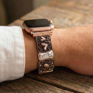 Puede incluir: Apple Watch en oro rosa con una correa decorativa. La correa presenta eslabones metálicos ornamentados con diseños de corazón y la palabra "Faith" en escritura cursiva. El reloj está en una muñeca.