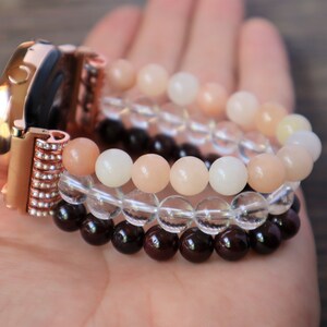 May include: A rose gold smartwatch band with a crystal-encrusted clasp. The band is adorned with three layers of beaded bracelets: a layer of clear quartz beads, a layer of peach-colored sunstone beads, and a layer of dark red garnet beads.