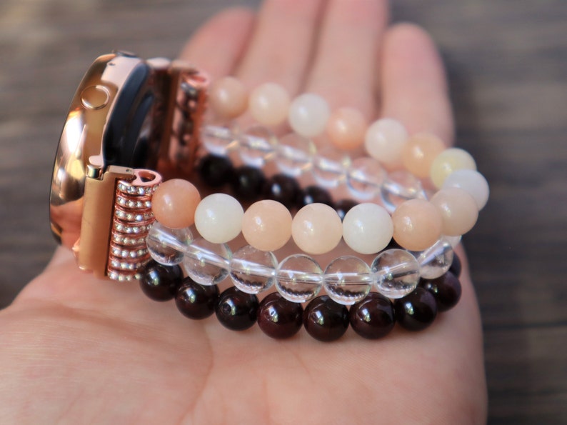 May include: A rose gold smartwatch band with a crystal-encrusted clasp. The band is adorned with three layers of beaded bracelets: a layer of clear quartz beads, a layer of peach-colored sunstone beads, and a layer of dark red garnet beads.