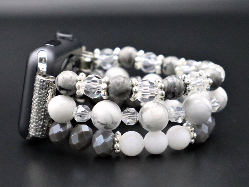 May include: A silver Apple Watch band made of grey and white beads. The band is made of three strands of beads, with the top strand being grey, the middle strand being white, and the bottom strand being grey.