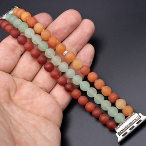 May include: A three-strand beaded Apple Watch band with green, orange, and brown beads. The band is made of smooth, round beads and has a silver metal clasp.