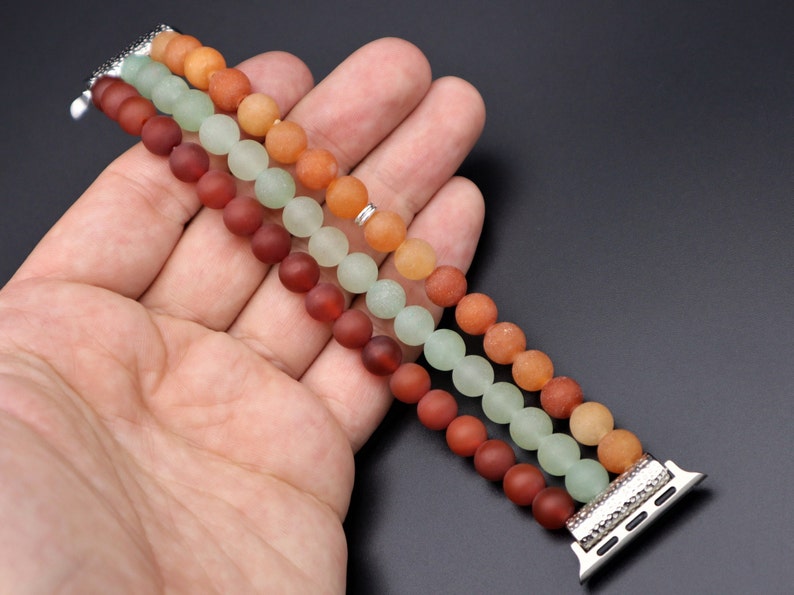 May include: A three-strand beaded Apple Watch band with green, orange, and brown beads. The band is made of smooth, round beads and has a silver metal clasp.