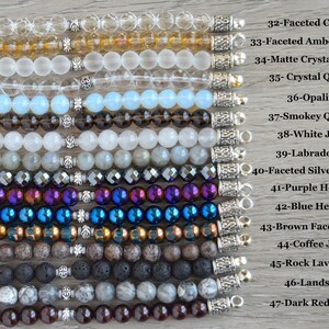 May include: A collection of 15 different gemstone bracelets with silver clasps. The bracelets are arranged vertically from top to bottom, with the names of each gemstone listed to the right of the image. The gemstones include faceted crystal, amber crystal, matte crystal quartz, crystal quartz, opalite, smokey quartz, white jade, labradorite, faceted silver hematite, purple hematite, blue hematite, brown faceted aqua, coffee jasper, rock lava beads, landscape, and dark red garnet.