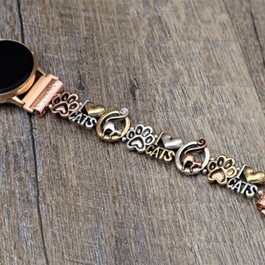 May include: A rose gold smartwatch band with charms featuring paw prints, cats, and hearts. The band is made of metal and has a polished finish.