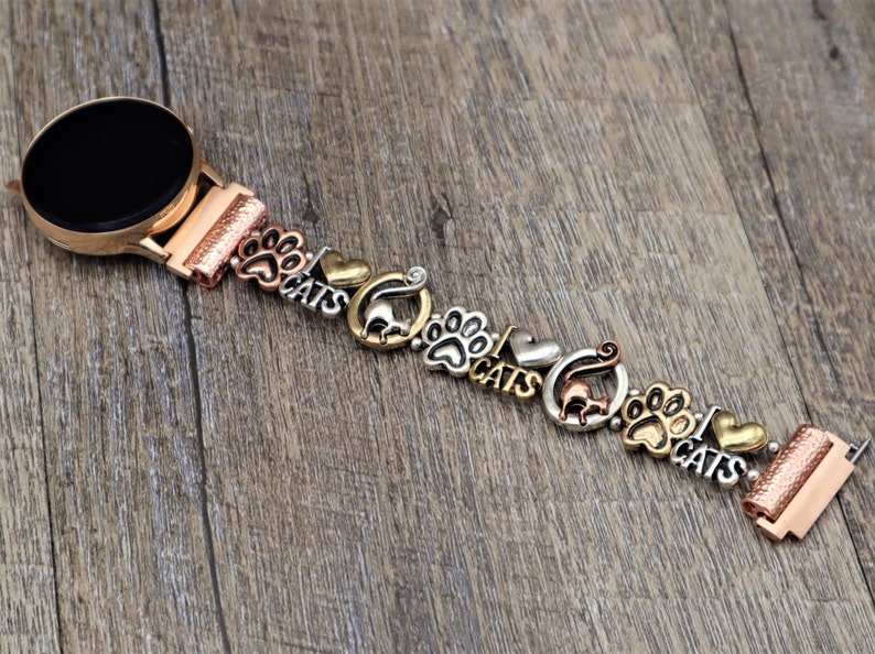 May include: A rose gold smartwatch band with charms featuring paw prints, cats, and hearts. The band is made of metal and has a polished finish.
