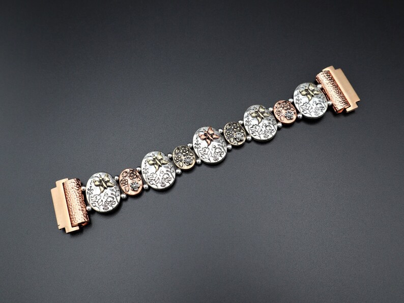 Pode incluir: Um bracelete de prata e cobre com um design floral e um fecho de encaixe. O bracelete &eacute; feito de metal e tem uma textura martelada.