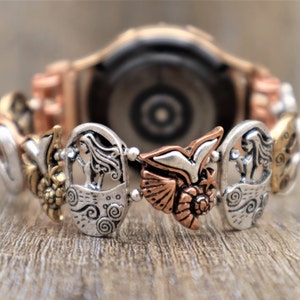 May include: A silver and copper watch band with a variety of charms, including a bird, a woman, and a flower. The watch band is designed to fit a smartwatch.