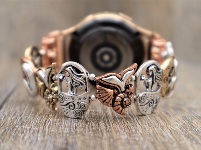 May include: A silver and copper watch band with a variety of charms, including a bird, a woman, and a flower. The watch band is designed to fit a smartwatch.
