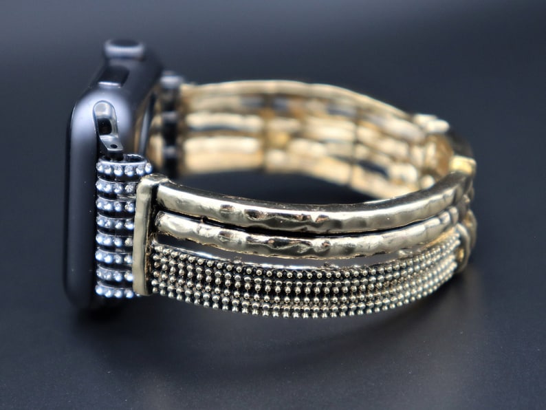 May include: A gold-toned watch band with a textured, hammered metal design. The band has a layered look with a row of small beads at the bottom.