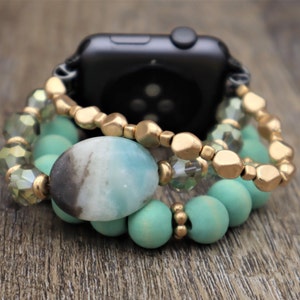 May include: A gold and turquoise beaded watch band with a large oval green and black gemstone. The band is made of multiple strands of beads, including gold, turquoise, and clear glass beads.