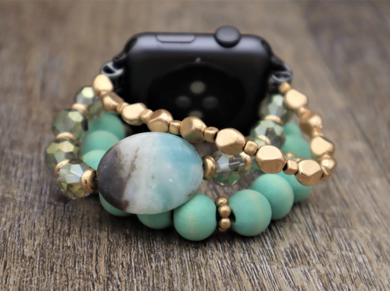 May include: A gold and turquoise beaded watch band with a large oval green and black gemstone. The band is made of multiple strands of beads, including gold, turquoise, and clear glass beads.