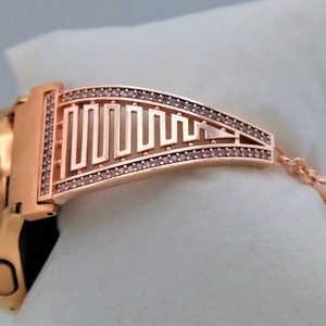 May include: Rose gold smartwatch band with a geometric design and clear rhinestones. The band has a clasp closure.