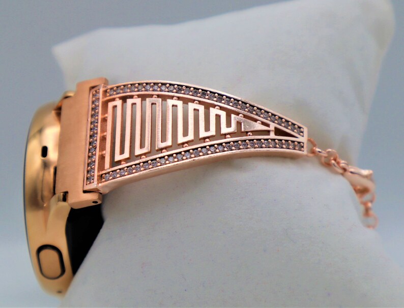 May include: Rose gold smartwatch band with a geometric design and clear rhinestones. The band has a clasp closure.