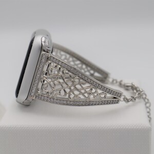 May include: A silver watch band with a decorative, intricate design featuring a chain link closure. The band is adorned with small, clear rhinestones.