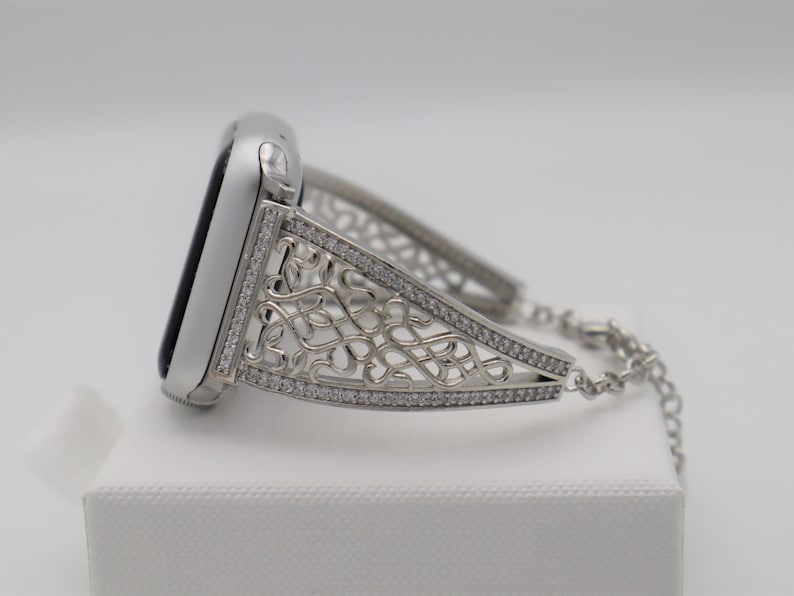 May include: A silver watch band with a decorative, intricate design featuring a chain link closure. The band is adorned with small, clear rhinestones.