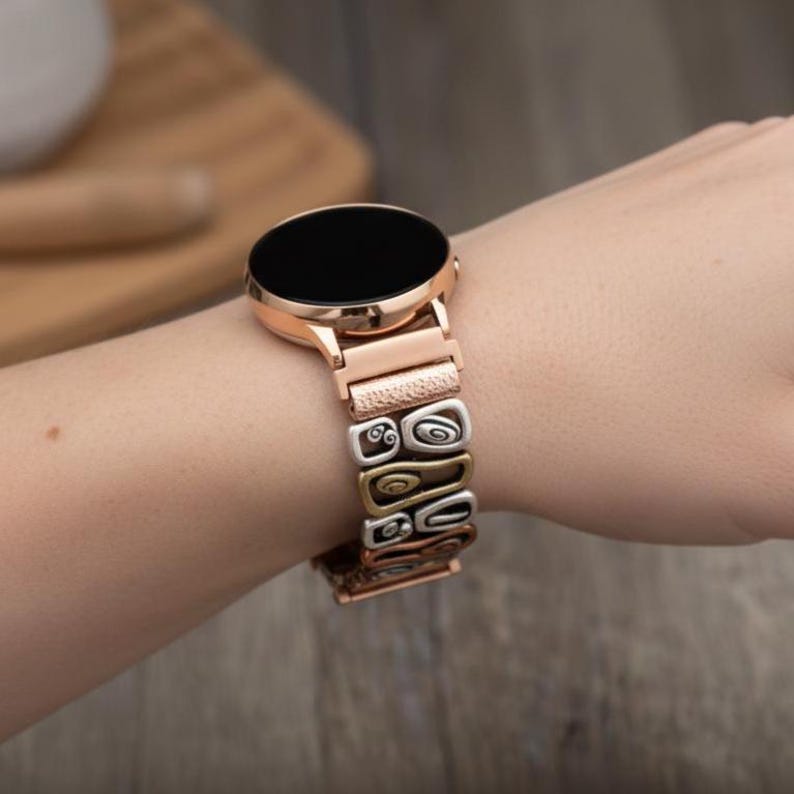 May include: Rose gold smartwatch with a black screen and a decorative band. The band features a mix of gold, silver, and bronze-colored metal links with an abstract design. The watch is on a wrist.