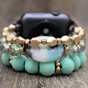 May include: A smartwatch with a turquoise and gold beaded band. The band is made of wooden beads, crystal beads, and gold-toned metal beads.