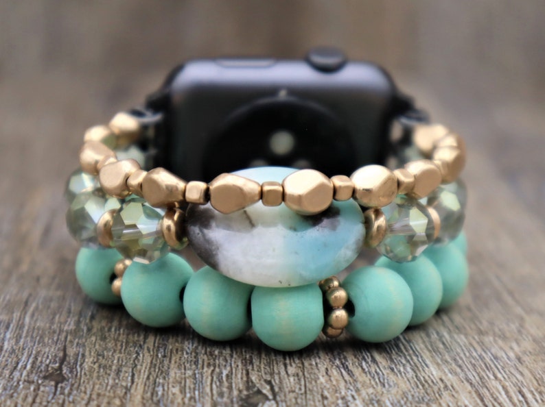 May include: A smartwatch with a turquoise and gold beaded band. The band is made of wooden beads, crystal beads, and gold-toned metal beads.