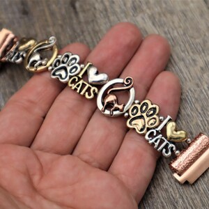May include: A silver and gold charm bracelet with cat-themed charms, including paw prints, hearts, and the words "I Love Cats".