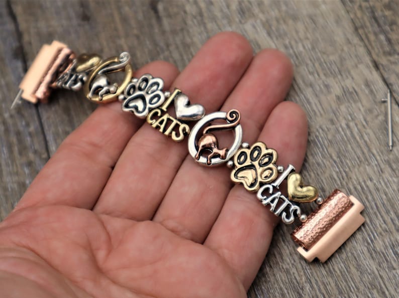 May include: A silver and gold charm bracelet with cat-themed charms, including paw prints, hearts, and the words "I Love Cats".