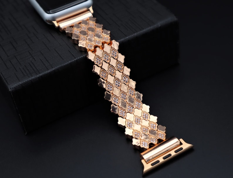 May include: A rose gold Apple Watch band with a diamond-like pattern. The band is made of metal and has a clasp closure.