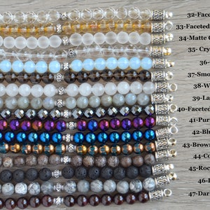 May include: A close-up of 15 different gemstone bracelets with silver clasps. The bracelets are arranged vertically, with the top bracelet being a clear faceted crystal and the bottom bracelet being a dark red garnet. The bracelets are labeled with their corresponding gemstone names.