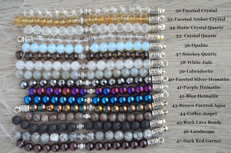 May include: A close-up of 15 different gemstone bracelets with silver clasps. The bracelets are arranged vertically, with the top bracelet being a clear faceted crystal and the bottom bracelet being a dark red garnet. The bracelets are labeled with their corresponding gemstone names.