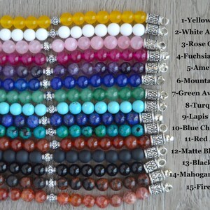 May include: Fifteen rows of gemstone bracelets with silver clasps. Each row features a different type of gemstone, including yellow agate, white alabaster, rose quartz, fuchsia quartz, amethyst, mountain jade, green aventurine, turquoise, lapis lazuli, blue chrysocolla, red agate, matte black onyx, black onyx, mahogany obsidian, and fire agate.