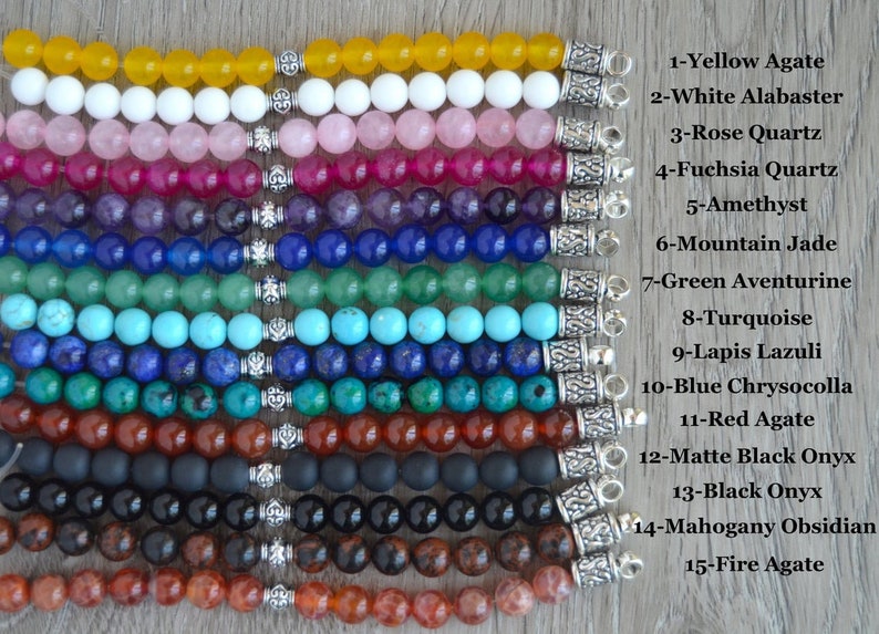 May include: Fifteen rows of gemstone bracelets with silver clasps. Each row features a different type of gemstone, including yellow agate, white alabaster, rose quartz, fuchsia quartz, amethyst, mountain jade, green aventurine, turquoise, lapis lazuli, blue chrysocolla, red agate, matte black onyx, black onyx, mahogany obsidian, and fire agate.