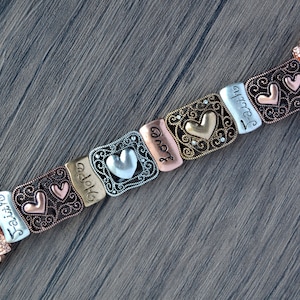 Puede incluir: Una correa de Apple Watch con eslabones met&aacute;licos decorativos. Los eslabones son de varios colores, incluyendo plata, bronce y oro rosa, y presentan dise&ntilde;os de coraz&oacute;n y las palabras "Faith", "Hope" y "Love".
