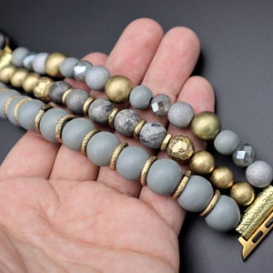 May include: A gray and gold beaded Apple Watch band with a three-row design. The band features gray, gold, and silver beads.
