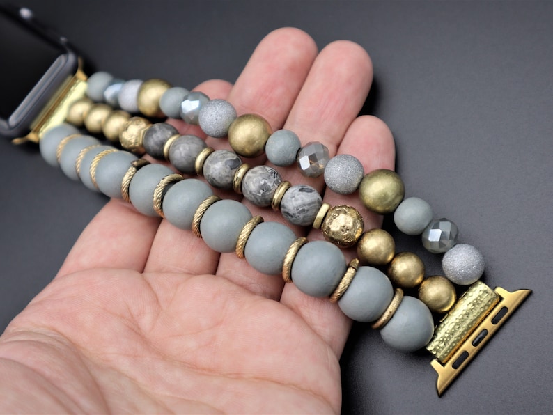 May include: A gray and gold beaded Apple Watch band with a three-row design. The band features gray, gold, and silver beads.