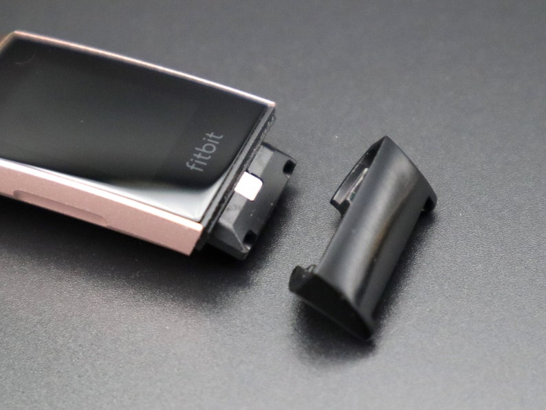 Puede incluir: Un reloj inteligente Fitbit de color rosa dorado con la palabra "fitbit" impresa en la esfera. El reloj est&aacute; separado de su banda negra.