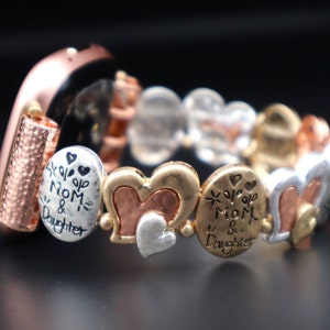 Può includere: Un braccialetto elasticizzato con perline in metallo misto. Il braccialetto presenta perline a forma di cuore e ovali in tonalità argento, oro e rame. Alcune perline hanno incise le parole "Mom & Daughter". Il braccialetto è su una superficie nera.