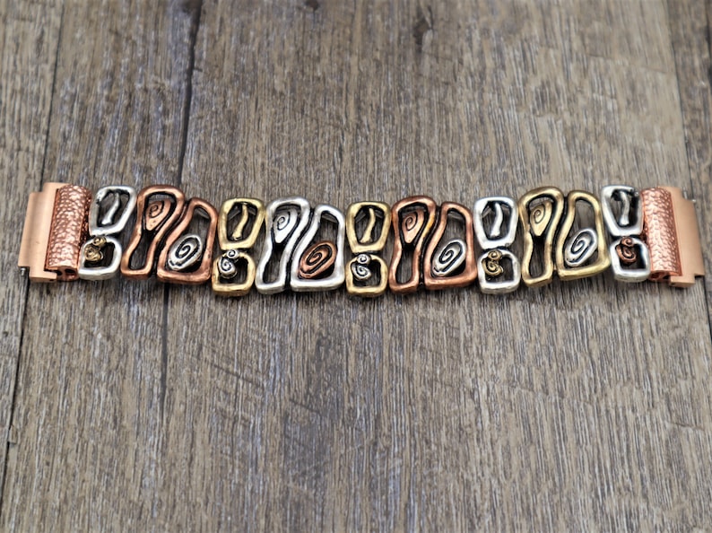 May include: A mixed-metal bracelet with a unique, abstract design. The bracelet features alternating sections of copper, silver, and gold-toned metal, with each section showcasing an open, swirling pattern. The clasp and end pieces are copper-colored.