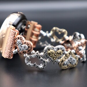 May include: A gold, silver, and rose gold watch band with a heart-shaped design. The band is made of metal and has a textured finish.