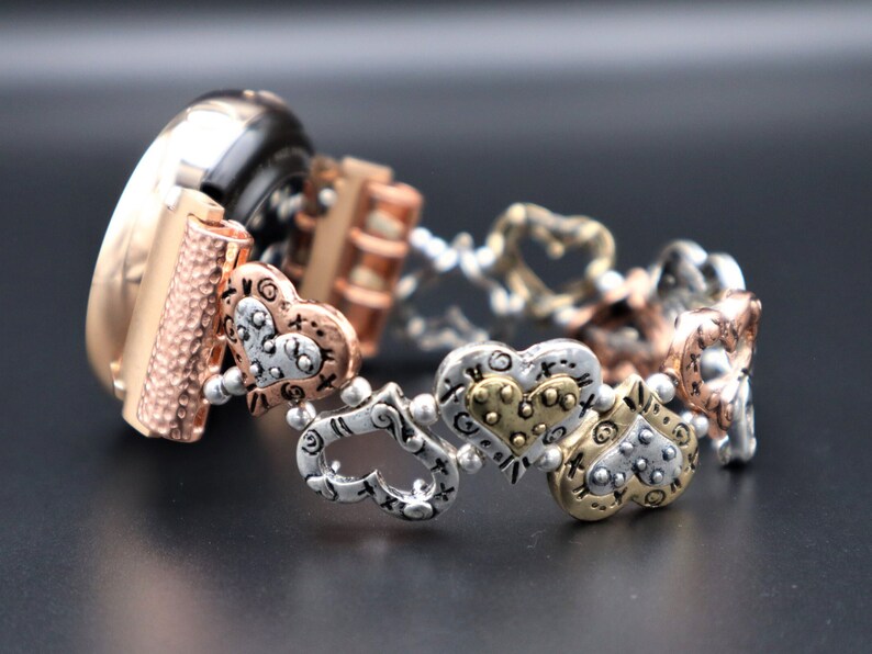 May include: A gold, silver, and rose gold watch band with a heart-shaped design. The band is made of metal and has a textured finish.