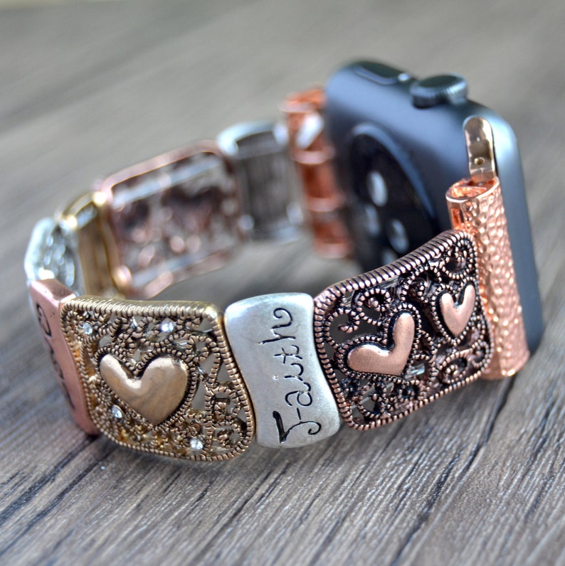 Puede incluir: Una correa de reloj Apple de metal multicolor, en plata, oro y cobre. La correa presenta filigranas decorativas, corazones y la palabra "Faith" en escritura cursiva. La correa se muestra sobre una superficie de madera.