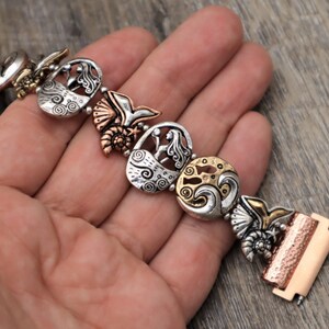 May include: A silver and copper bracelet with a mermaid, seashell, and wave design. The bracelet is made of metal and has a clasp closure.