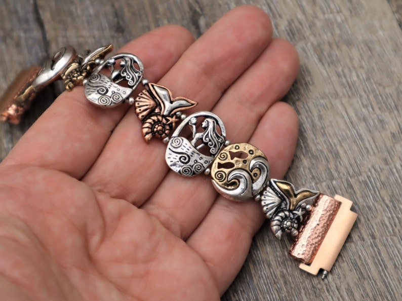 May include: A silver and copper bracelet with a mermaid, seashell, and wave design. The bracelet is made of metal and has a clasp closure.