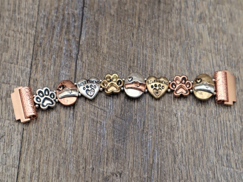 Puede incluir: Pulsera de metal multicolor con huellas de patas, corazones y abalorios circulares. La pulsera incluye las palabras "BEST FRIEND" en abalorios en forma de coraz&oacute;n. El cierre y algunos abalorios tienen un acabado cobrizo.