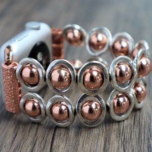 Puede incluir: Una pulsera de Apple Watch de plata y oro rosa con un diseño de cuentas. La pulsera está hecha de cuentas de metal con un diseño circular y un acabado en oro rosa.