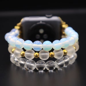 K&ouml;nnte beinhalten: Ein dreireihiges Perlenarmband f&uuml;r eine Uhr mit einem goldenen Verschluss. Die obere Reihe besteht aus undurchsichtigen wei&szlig;en Perlen, die mittlere Reihe aus klaren Perlen mit goldenen Akzenten und die untere Reihe aus klaren Perlen.