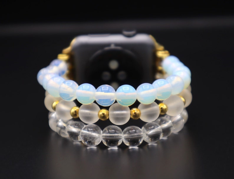 K&ouml;nnte beinhalten: Ein dreireihiges Perlenarmband f&uuml;r eine Uhr mit einem goldenen Verschluss. Die obere Reihe besteht aus undurchsichtigen wei&szlig;en Perlen, die mittlere Reihe aus klaren Perlen mit goldenen Akzenten und die untere Reihe aus klaren Perlen.