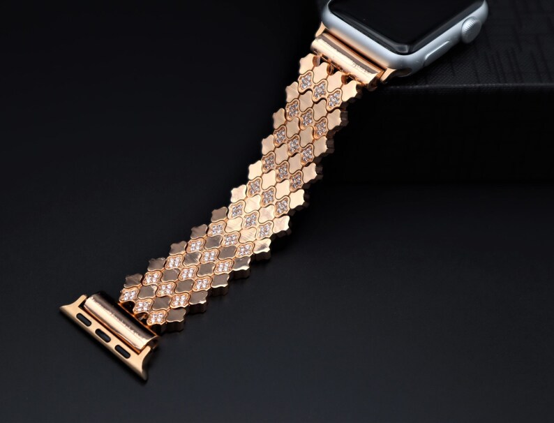 May include: A gold-tone Apple Watch band with a diamond-like pattern. The band is made of metal links and has a clasp closure.