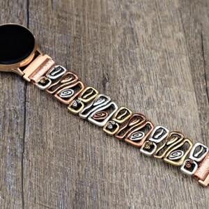 May include: A rose gold smartwatch with a multi-tone metal band. The band features a repeating pattern of copper, silver, and gold-colored links. The watch face is black and round, and the band is connected to the watch with a rose gold clasp.