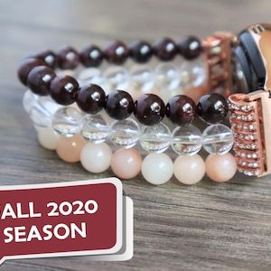 Puede incluir: Un smartwatch de oro rosa con una banda adornada con cristales. El reloj está adornado con tres pulseras de cuentas superpuestas: granate rojo oscuro, cuarzo transparente y piedra lunar rosa claro.