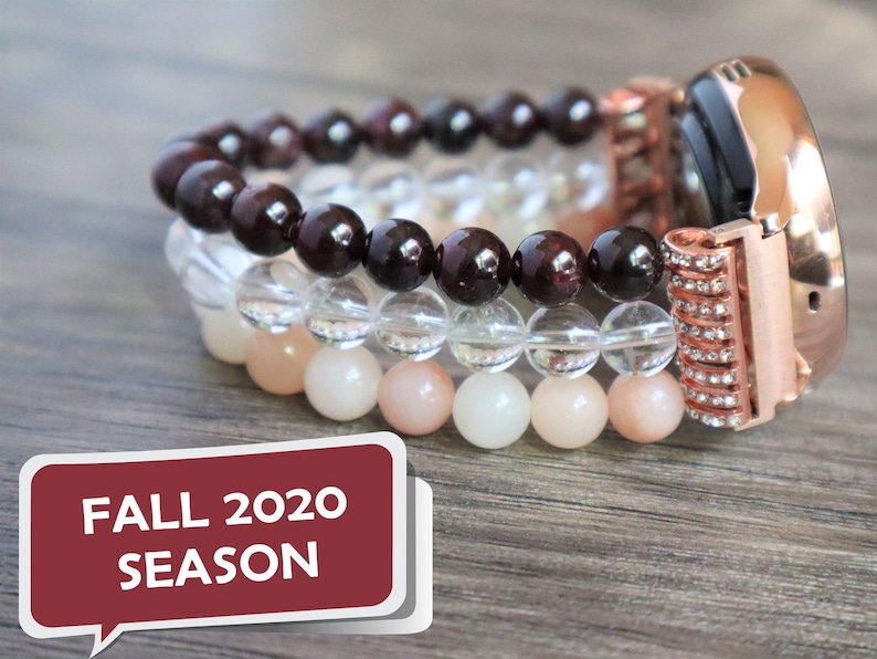 May include: A rose gold smartwatch with a crystal-encrusted band. The watch is adorned with three layered beaded bracelets: dark red garnet, clear quartz, and light pink moonstone.