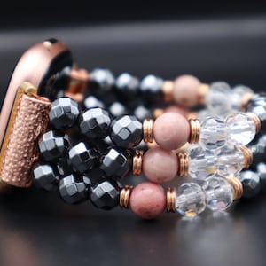 Può includere: Uno smartwatch color oro rosa con un cinturino di perline. Il cinturino è composto da tre file di perline: ematite nera, rosa e cristallo trasparente.